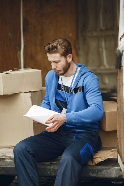 A young man with light brown hair and a beard, dressed in a blue hoodie and dark blue work trousers, is seated on the edge of a moving van's open cargo area, surrounded by cardboard boxes and packing materials. He is holding and reading a document, possibly related to the home relocation or moving process. Behind him, there are additional cardboard boxes, some stacked, and protective packing blankets. The setting appears to be a partially enclosed space or driveway, with wooden paneling or fencing visible in the background. The lighting is natural, illuminating the scene clearly. This image illustrates the preparatory stages of a professional removals service, such as those offered by Man with Van Hounslow, featuring packing, documentation review, and transportation planning in the context of a household move.