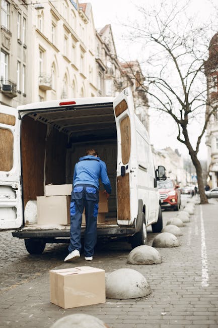A man wearing a blue jacket and dark trousers is seen loading or unloading packing boxes from the back of a white moving van parked on a cobblestone street. The van's rear doors are open, revealing empty wooden interior walls and the interior space designated for transporting furniture or household items during a home relocation. Several cardboard boxes, including one placed on the ground nearby, are part of the items being moved. The street is lined with multi-story buildings with classic architectural features, and there are large concrete bollards along the edge of the sidewalk preventing vehicle encroachment. Bare trees with thin branches suggest a cold season. The lighting is natural daylight, and the scene captures the logistics involved in packing and moving, indicative of professional removals services like those offered by Man with Van Hounslow.