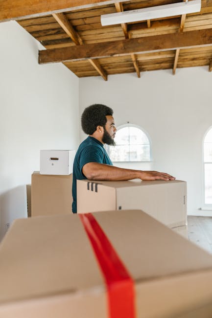A man with a beard and curly hair, dressed in a teal shirt, is standing inside an empty room during a home relocation process. He is positioned behind a stack of large cardboard boxes, with one box pl