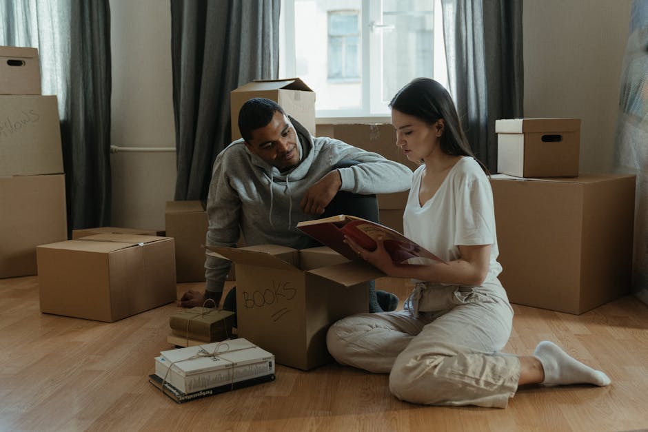 A man and a woman are inside a room with wooden flooring, surrounded by various cardboard boxes used for a house relocation. The woman, sitting cross-legged on the floor, is holding an open book and appears to be reading, while the man, kneeling beside her, is leaning on a cardboard box labeled 'BOOKS' with his arm resting on it. Some of the boxes are sealed with tape, and others are open, revealing packing materials such as paper and smaller boxes stacked on top. In the background, there are additional cardboard boxes of different sizes positioned against a wall with a window, partly covered by dark curtains. The room has natural light coming through the window, and the scene captures the process of packing and preparing for furniture transport, with minimal furniture visible, emphasizing the packing and moving logistics handled by a professional removal service like Man with Van Hounslow.