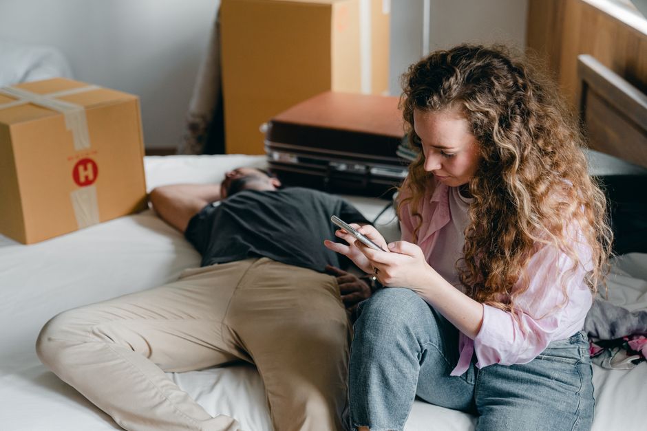 A woman with long, curly hair and wearing a pink shirt sits on the edge of a bed, holding and looking at a smartphone. Behind her, a man with short dark hair and dressed in a black t-shirt and beige trousers lies on the bed on his side, facing away from the woman and resting with his arm over his head. The room features a wooden headboard, and several cardboard boxes sealed with packing tape are positioned on the bed and nearby, indicating packing or a home relocation process. To the side, there are stacked suitcases and a pile of cardboard boxes, some with visible labels, inside a room with natural lighting. The scene captures an indoor moving or packing situation, with visible packing materials and furniture suggesting an ongoing house removal or logistics task managed by [COMPANY_NAME], associated with home moving services in Hounslow.