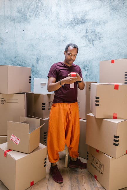 A man and a woman are inside a room with wooden flooring, surrounded by various cardboard boxes used for a house relocation. The woman, sitting cross-legged on the floor, is holding an open book and appears to be reading, while the man, kneeling beside her, is leaning on a cardboard box labeled 'BOOKS' with his arm resting on it. Some of the boxes are sealed with tape, and others are open, revealing packing materials such as paper and smaller boxes stacked on top. In the background, there are additional cardboard boxes of different sizes positioned against a wall with a window, partly covered by dark curtains. The room has natural light coming through the window, and the scene captures the process of packing and preparing for furniture transport, with minimal furniture visible, emphasizing the packing and moving logistics handled by a professional removal service like Man with Van Hounslow.