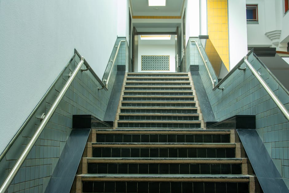A photograph of a staircase inside a residential building, showing wooden steps with black tiles on the risers and sides. The stairway is flanked by stainless steel handrails attached to light-colored tiled walls. At the top of the stairs, there is a door with a vented panel below it, leading to an upper floor. The area is well-lit with natural light coming from a window or opening at the top of the staircase. This interior view illustrates part of the stair access point commonly encountered during home relocations or furniture transport, relevant to house removals and packing services offered by Man with Van Hounslow.