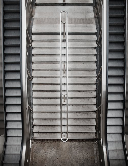 A top-down view of a concrete staircase inside a building, with metal handrails on either side. The staircase has wide, evenly spaced concrete steps leading downward, with a vertical metal safety gate installed at the top, featuring a handle oriented vertically. To the left and right of the staircase are dark metal escalator steps, indicating an escalator adjacent to the staircase. The surrounding area has light-colored tiled flooring at the top of the stairs, and the photo captures the clean, structured environment typical in residential or commercial building corridors, supporting efficient home relocation or furniture transport in stair-access scenarios as part of professional removals services provided by Man with Van Hounslow.