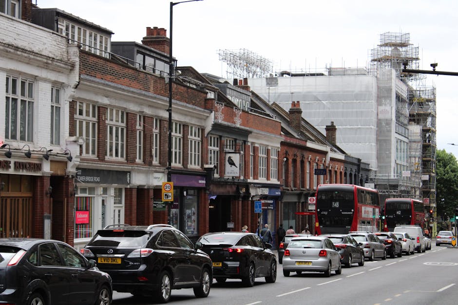 A narrow street flanked by multi-storey residential buildings with aged facades and exposed brickwork, situated in an urban area of Hounslow. Several parked cars, including a silver vehicle and an orange-striped van, line both sides of the pavement. The street appears confined, with overhead wires running between the buildings. In the background, a hillside densely populated with additional residential structures extends upward, indicating a hilly terrain typical of some housing areas. Lighting is natural with overcast skies, and there are no visible people or moving objects in the scene, which relates to the planning phase of home relocation or furniture transport via professional removal services such as Man with Van Hounslow, who specialise in house removals and moving logistics in tight urban environments.