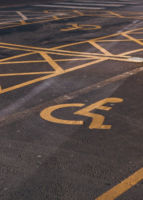A close-up view of a parking lot surface showing multiple yellow-painted disabled parking bay markings, including the wheelchair symbol and diagonal lines, on dark asphalt. The markings are positioned near the entrance of a property, with a section of the pavement visible in the foreground. The scene is outdoors, with no vehicles or moving objects present, and the area appears well-maintained. This setting is relevant to house removals and moving services provided by Man with Van Hounslow, highlighting convenient access points for loading or unloading during a property relocation near Osterley Park, with attention to proper parking arrangements and access considerations.