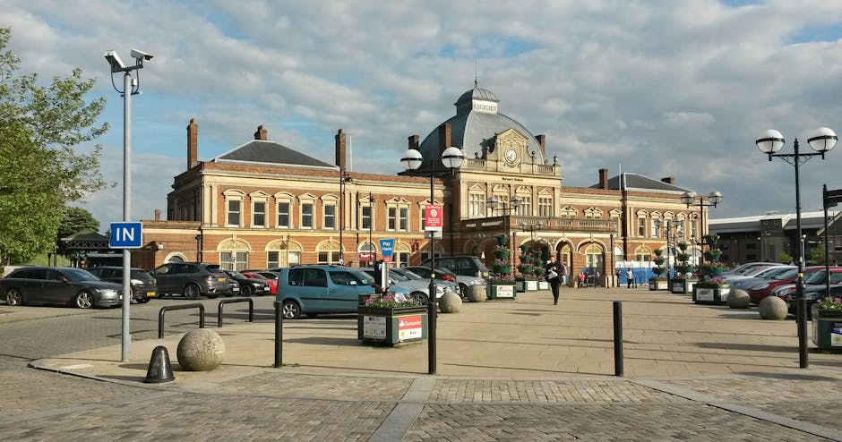 A large, historic train station building constructed from red brick with cream decorative features, situated in a busy town square. In the foreground, there is a paved area with several parked cars, including small hatchbacks and sedans, along with black metal barriers and large spherical bollards lining the pavement. A blue sign marked 'IN' indicates vehicle entry, and a few pedestrians are walking nearby. Tall street-lamp posts with multiple spherical lamps are positioned around the square, some decorated with hanging flower baskets. The sky is partly cloudy with patches of sunlight illuminating the scene. This setting appears to be a town centre, where a professional house removals company like Man with Van Hounslow might operate to facilitate efficient furniture transport and home relocation services, highlighting the parking and access conditions near Osterley Park.