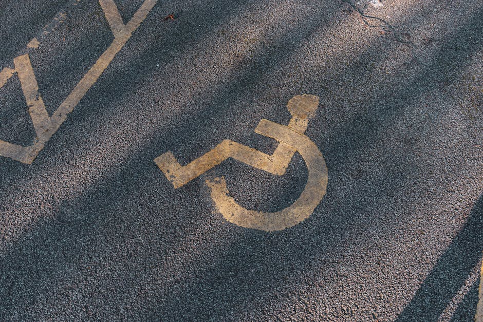 A close-up view of a parking lot surface showing multiple yellow-painted disabled parking bay markings, including the wheelchair symbol and diagonal lines, on dark asphalt. The markings are positioned near the entrance of a property, with a section of the pavement visible in the foreground. The scene is outdoors, with no vehicles or moving objects present, and the area appears well-maintained. This setting is relevant to house removals and moving services provided by Man with Van Hounslow, highlighting convenient access points for loading or unloading during a property relocation near Osterley Park, with attention to proper parking arrangements and access considerations.