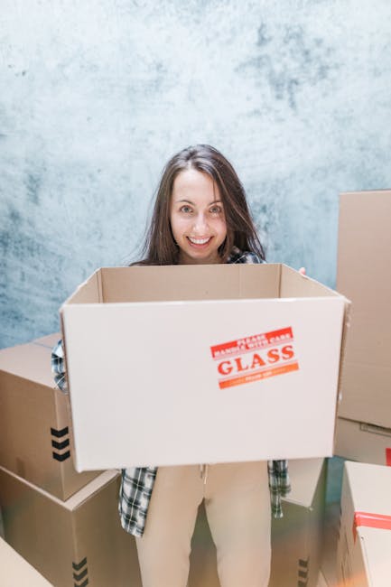 A young woman with curly black hair and a bright smile is sitting on the wooden floor inside a room, surrounded by numerous cardboard boxes of varying sizes, some sealed with red or black tape and oth