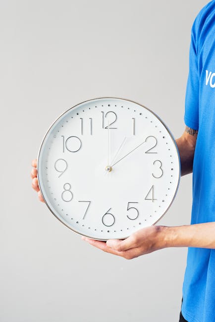 A person wearing a blue T-shirt is holding a large, round wall clock with a white face and black numerals. The clock shows a time just after 12:00, with the minute hand pointing slightly past the 12 and the second hand just after the 2-minute mark. The individual is gripping the clock on its sides, with their right hand visible. The background is plain and light gray, creating a neutral setting. This image illustrates the concept of time management during home relocation or furniture transport, relevant to [PAGE_TITLE], with [COMPANY_NAME] providing removals services. The scene focuses on the clock as a key element, emphasizing the importance of punctuality in moving logistics and packing during a same-day move.