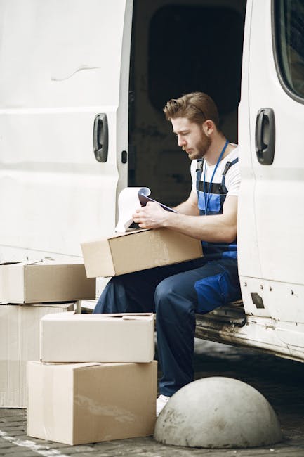 A man wearing a blue and white striped shirt and dark blue overalls is sitting on the edge of a white van with open sliding doors during a home relocation process. He is holding a clipboard and examining documents or a packing list, with several cardboard boxes of varying sizes placed around him on the ground. Some boxes are stacked, and others are open, revealing contents wrapped in protective packaging material. The van is parked on a paved surface, and a concrete bollard is visible nearby. The background suggests an outdoor setting, possibly a driveway or parking area, with natural lighting illuminating the scene. This image depicts the loading or packing phase of furniture transport and moving services offered by Man with Van Hounslow, focusing on careful handling and organization of packed items in preparation for delivery in a house removal or furniture transport context.