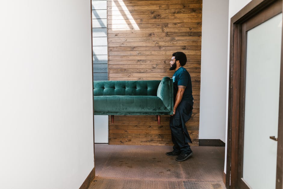 A man with dark curly hair and a beard, wearing a dark blue uniform, is inside a room with wooden paneling on one wall and white painted walls on the other. He is carrying a large, green, velvet-upholstered sofa with wooden legs, hoisted vertically with the cushions facing inward. The sofa is partially inside the room, positioned vertically, and it appears to be in the process of being moved through a doorway. The room has a brown carpeted floor, and daylight streams in from an unseen window or light source, creating shadows on the walls and floor. This scene captures the careful lifting and positioning involved in home relocation, likely as part of a furniture transport and packing process by Man with Van Hounslow, specializing in small flat removals and house moving services on Staines Road, Hounslow.